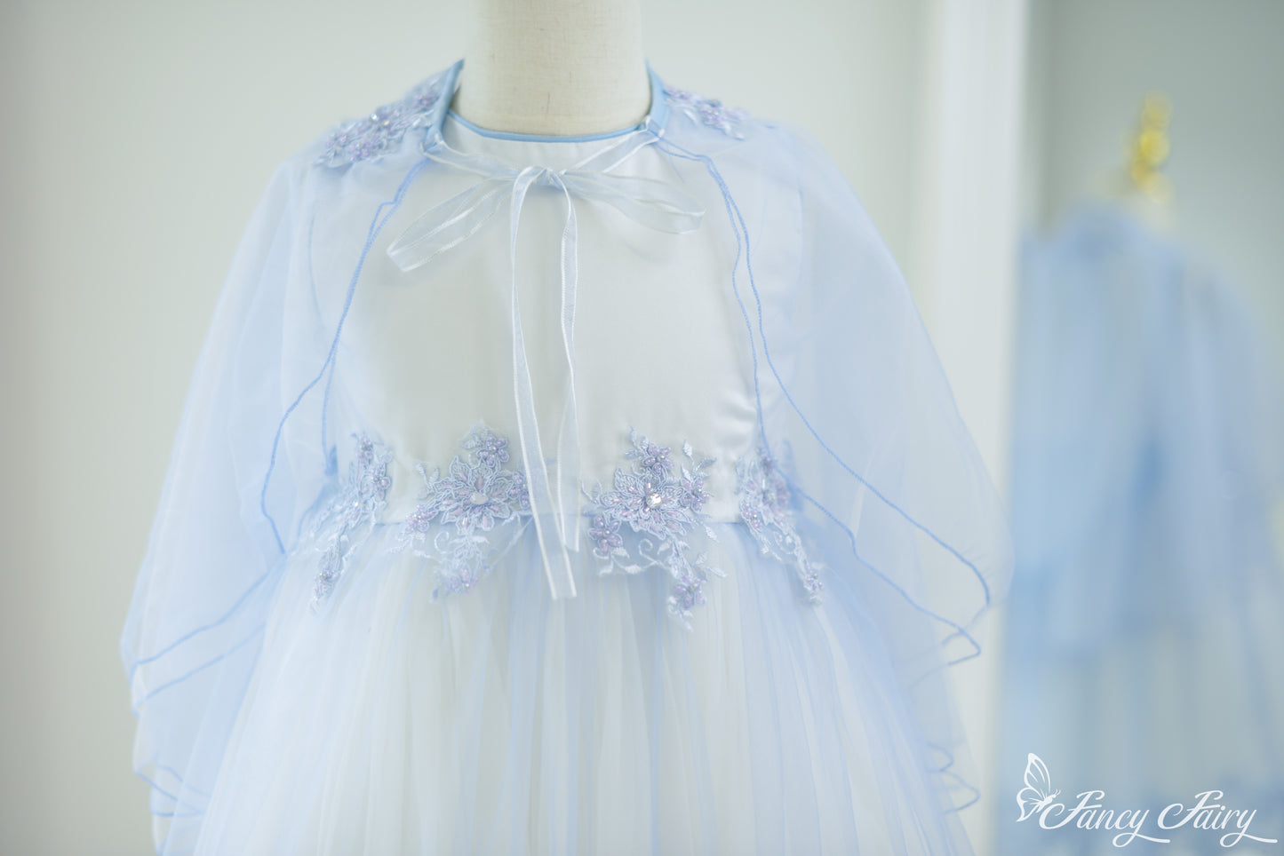 Front view of light blue A-line girls' dress with sheer cape and floral embroidery.